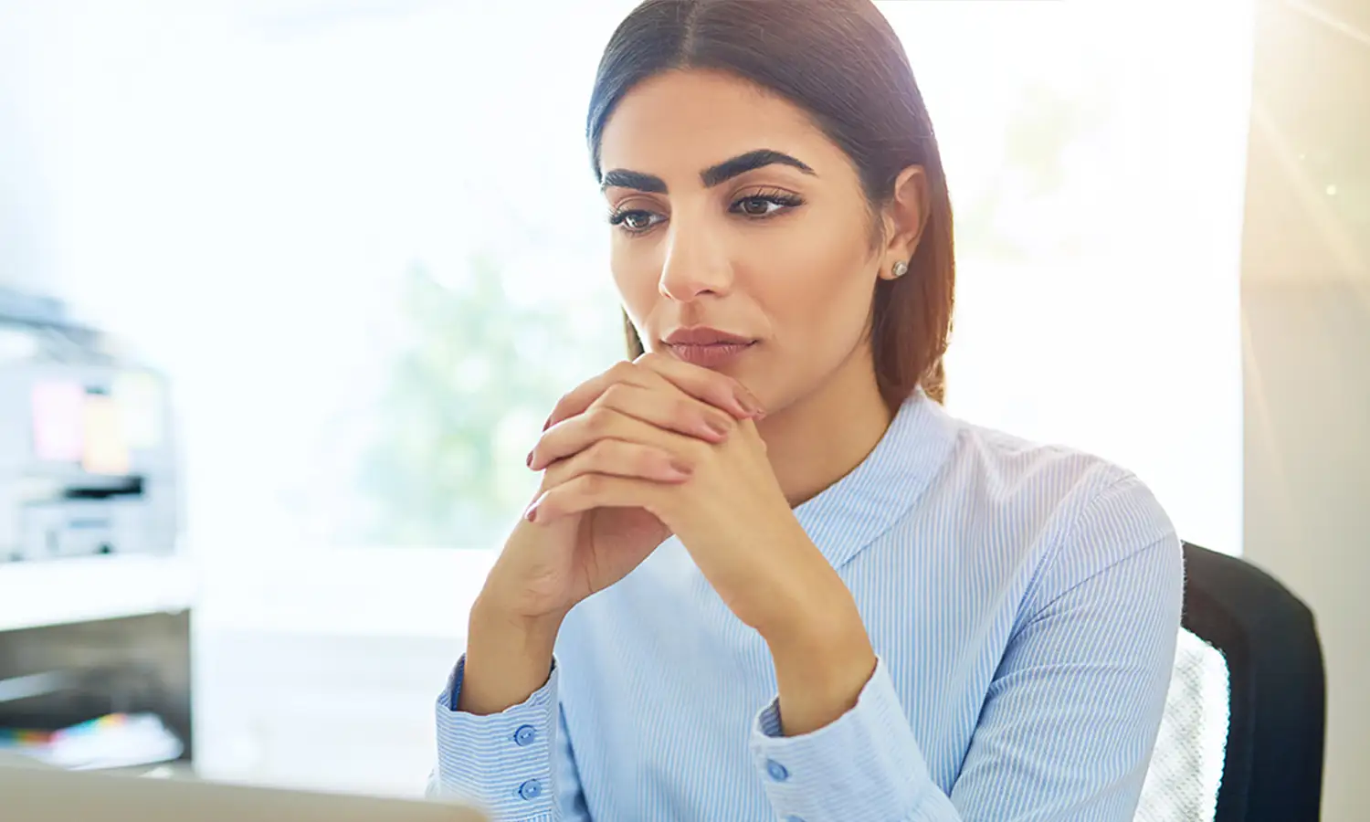 Woman sitting at a desk with hands clasped, appearing focused while looking at a computer screen in a bright office.