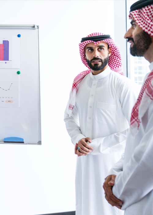 Two Saudi men in thobes discuss business beside charts in a modern office.